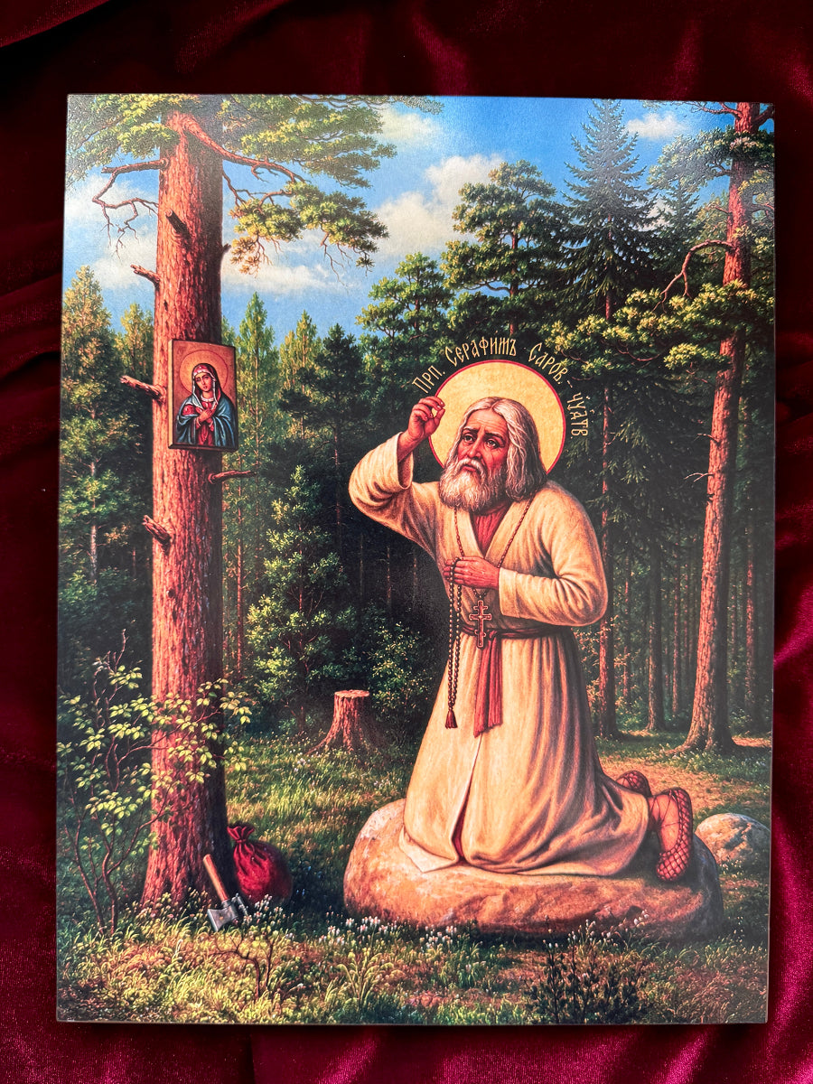 A rectangular religious print or icon depicting a kneeling saint Seraphim in a forest beside a tree with a small icon of the Virgin Mary, placed on a dark red fabric background.