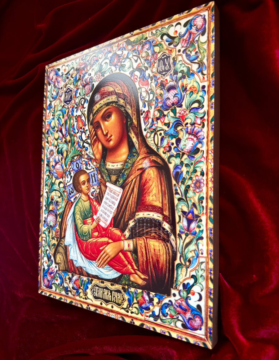 An ornate religious icon painting of the Virgin Mary holding the infant Jesus, shown at an angle against a rich red velvet background. The icon features intricate multicolor floral patterns and detailed gold, blue, green, and red elements.