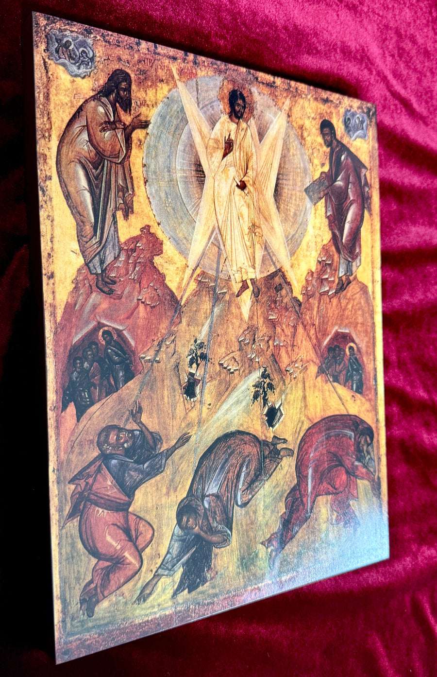 An Eastern Orthodox icon painting depicting the Transfiguration, mounted on a wooden panel and displayed on a red velvet fabric background.